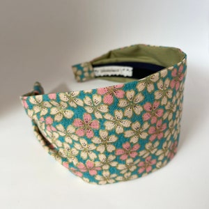 May include: A teal blue headband with a floral pattern featuring pink and white flowers. The headband is made of fabric and has a knotted design.