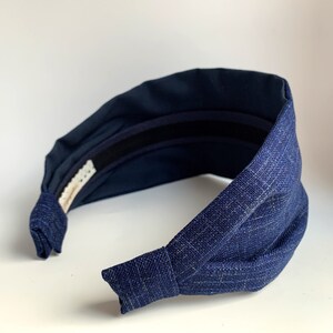 May include: A dark blue fabric headband with a knotted design. The headband is made of a textured fabric and has a black elastic band on the inside.