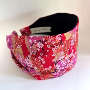 May include: Red headband with a gold and pink floral pattern. The headband is made of fabric and has a black elastic band.