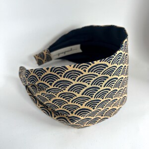May include: A black and gold headband with a wave pattern. The headband is made of fabric and is designed to be worn around the head.