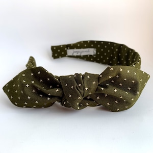 May include: A green headband with a white cross pattern and a large bow. The headband is made of fabric and is designed to be worn around the head.