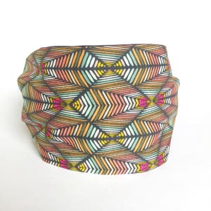 May include: A colorful headband with a geometric pattern. The fabric features a repeating design of stylized arrows in shades of brown, orange, pink, green, yellow, and white on a black background. The headband is folded and appears soft.