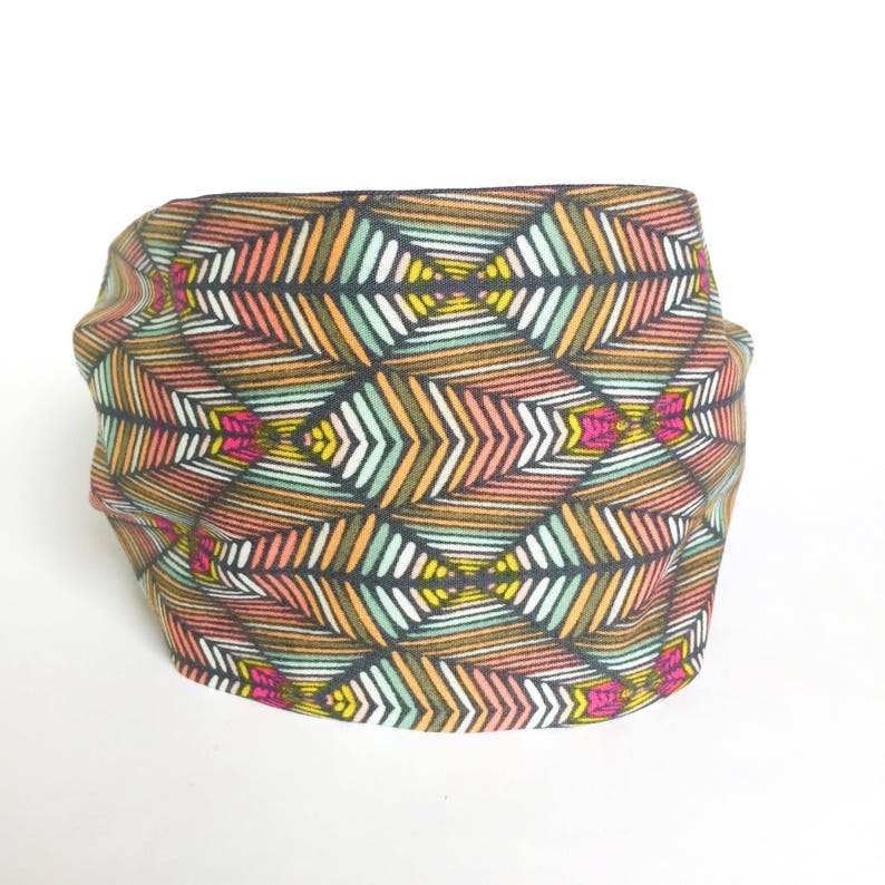 May include: A colorful headband with a geometric pattern. The fabric features a repeating design of stylized arrows in shades of brown, orange, pink, green, yellow, and white on a black background. The headband is folded and appears soft.