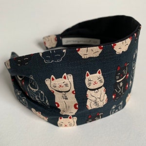 May include: A black headband with a repeating pattern of white lucky cats with red accents on a dark blue background.