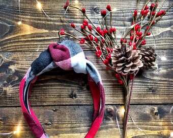 May include: A red, black, and white plaid headband with a knotted bow. The headband is on a wooden background with red berries and pine cones.