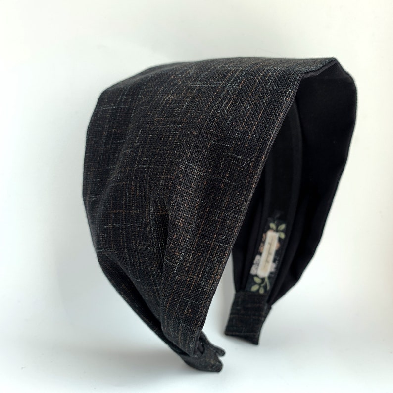 K&ouml;nnte beinhalten: Ein schwarzes Stoffhaarband mit einem geknoteten Design. Das Haarband ist aus einem strukturierten Stoff mit einem dezenten Glanz.