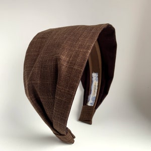 May include: A brown fabric headband with a knotted design. The headband is made of a textured fabric and has a brown elastic band.