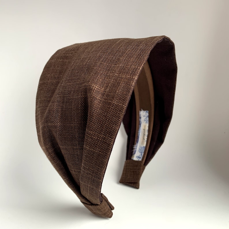 May include: A brown fabric headband with a knotted design. The headband is made of a textured fabric and has a brown elastic band.