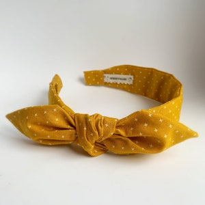 May include: Mustard yellow headband with a knotted bow. The fabric features a white cross pattern. The interior lining is a solid mustard yellow. The label reads "JERSEYMAID".