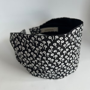 May include: Black headband with a white pattern of small circles. The headband is made of fabric and is designed to be worn around the head.