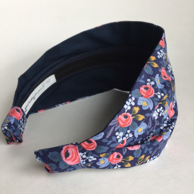 May include: A wide headband with a floral pattern. The headband features a navy blue background with a repeating pattern of pink roses, blue flowers, and green leaves. The interior is a solid navy blue, and the label reads "jerseymaid".