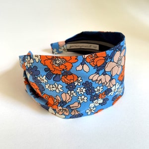 May include: A blue headband with a floral pattern featuring orange, pink, and white flowers. The headband is made of fabric and has a navy blue elastic band.