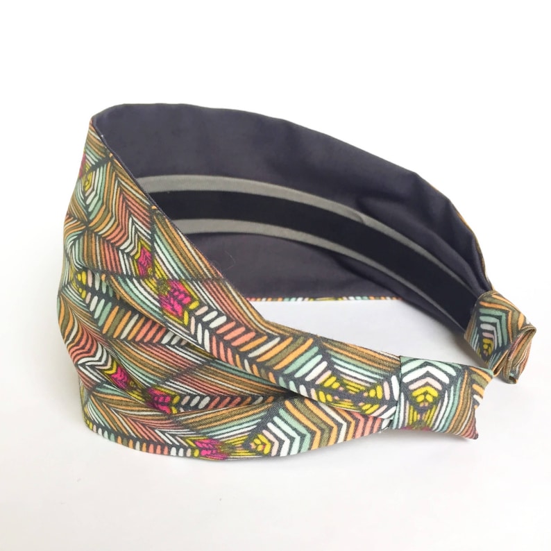 May include: A wide headband with a geometric pattern in shades of orange, yellow, green, and pink. The interior is a solid dark gray, with two gray stripes. The headband is made of fabric.