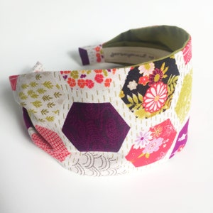 May include: A patterned headband with a floral and geometric design. The fabric has a white base with colourful hexagonal shapes in purple, pink, and yellow, and floral patterns. The interior is green, and the label reads "jerseymade".