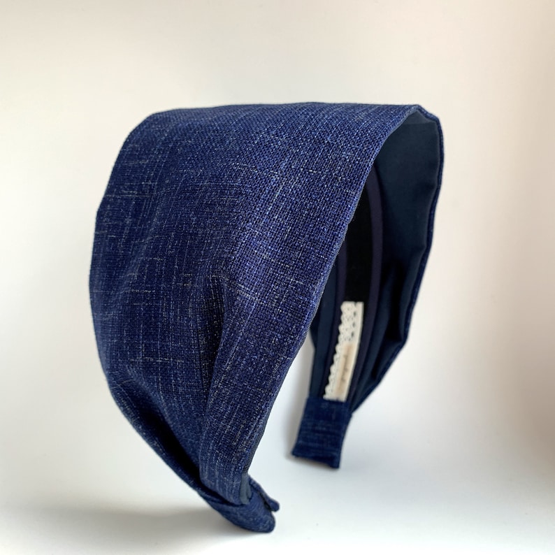 May include: A dark blue fabric headband with a knotted design. The headband is made of a textured fabric and has a simple, elegant style.