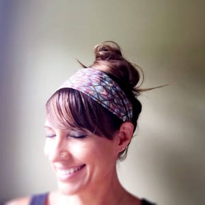 May include: A colorful patterned headband in shades of pink, purple, and blue is worn on the head. The headband is wrapped around the forehead and is paired with a hairstyle that includes bangs and a bun. The person is smiling.