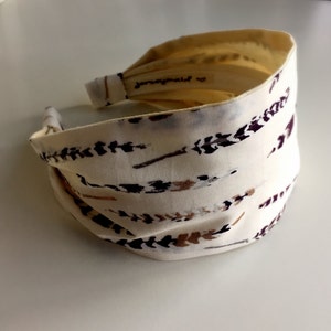 May include: A white fabric headband with a brown and purple feather pattern. The headband is folded and has the text "Jennymaid" on the inside.