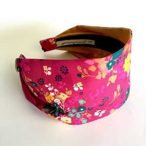 May include: A pink headband with a floral pattern. The headband is made of fabric and has a knotted design. The headband has a label that says "jerseymaid".