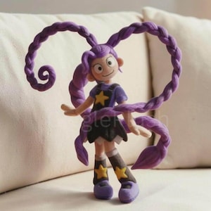 Op de afbeelding: Een klein figuurtje van een personage met lang, paars gevlochten haar dat omhoog krult. Het personage draagt een paars shirt met een gele ster, een zwarte rok en bijpassende laarzen met gele sterren. Het figuurtje heeft een lichte huidskleur.