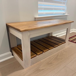 Peut inclure: Un banc rectangulaire en bois doté d'un plateau au grain naturel et d'une étagère inférieure à lattes. La structure et les pieds présentent une finition blanchie, tandis que le plateau et les lattes de l'étagère sont teintés en brun moyen.