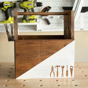 May include: A rectangular wooden tool caddy with a top handle, featuring a dark brown stained finish on one side and a white painted diagonal section on the other. The white area displays brown silhouettes of a pliers, hammer, wrench, screwdriver, and saw.