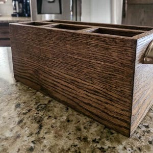 May include: A rectangular dark brown wooden organizer with a visible wood grain texture, featuring three open compartments on the top and a single brushed silver metal cup pull handle attached to the side of the box for easy handling.
