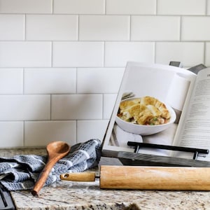 May include: A wooden rolling pin, a wooden spoon, and a grey and white plaid kitchen towel resting near an open cookbook on a black metal stand. The cookbook page displays a recipe for chicken pot pie with a photograph of the prepared dish.