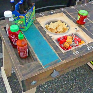 Könnte beinhalten: Ein Holztisch im Design eines Footballfeldes mit Yard-Markierungen, einer blauen Harzeinlage, einem integrierten Flaschenöffner und vertieften Fächern für eine Schüssel Tortilla-Chips, verschiedene Süßigkeiten, scharfe Soße und Bier.