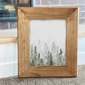 Könnte beinhalten: Ein rechteckiger Bilderrahmen aus Holz mit hellbrauner Oberfläche, der ein Aquarellbild eines Waldes enthält. Das Kunstwerk zeigt Schichten von immergrünen Bäumen in Grün- und Grautönen, die eine neblige, tiefenreiche Landschaftsszene bilden.