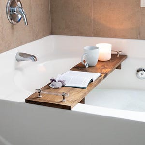 May include: A rectangular dark brown wooden bathtub tray featuring clear acrylic handles with silver-toned hardware. The tray holds an open book, a white ceramic mug with a tea bag tag, a glowing white candle, and a small cluster of raw purple crystals.