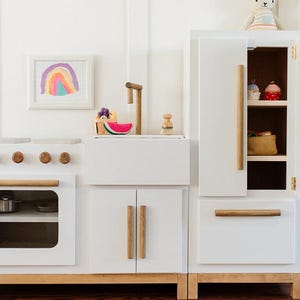 Op de afbeelding: Een witte houten speelkeuken met een fornuis met vier knoppen, een oven met een venster, een gootsteen met een houten kraan en een hoge kast met open deuren. De set is voorzien van handgrepen en accenten aan de onderkant van licht hout.