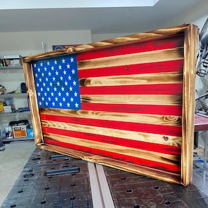 Puede incluir: Una pieza de arte mural rectangular de madera con un diseño de bandera estadounidense con acabado de madera quemada. La bandera tiene un cantón azul con estrellas blancas y franjas alternas rojas y de madera natural, todo dentro de un marco grueso teñido.
