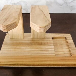 May include: A light-coloured wooden docking station featuring two elevated, angled arms designed for hanging items and a rectangular recessed tray on the base. The natural wood grain is visible throughout the smooth, polished surface of the stand.