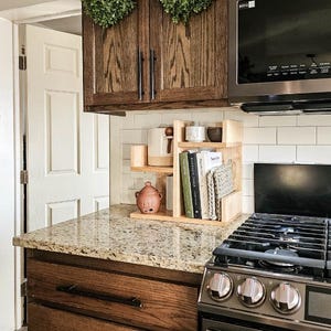 May include: A light-colored wooden countertop organizer with two tiers, holding a ceramic pitcher, a small terracotta pot, and several books. The organizer is positioned on a granite countertop next to a gas stove and beneath dark wood cabinets.