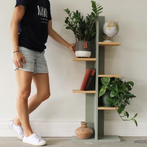 May include: A tall, sage green wooden shelving unit with five staggered light wood shelves. The unit holds a potted plant, a small globe, two books, a second potted plant, and a terracotta vase. The text WOODSHOP DIARIES is in the bottom right.
