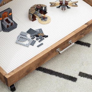 May include: A rectangular wooden rolling storage tray featuring a white building block base plate surface. The tray includes a silver metal handle on the front edge and small black wheels underneath, containing various plastic building block structures.