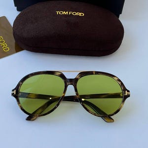 Può includere: Occhiali da sole aviatore con montatura tartarugata marrone, ponte in metallo dorato e lenti verde chiaro. Include una custodia rigida in finta pelle scamosciata marrone scuro con il testo dorato TOM FORD e una scatola rettangolare nera con TOM FORD.