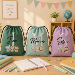 Puede incluir: Tres mochilas de cordón en verde oscuro, verde claro y lavanda, cada una con una inicial blanca grande, un nombre personalizado en letra cursiva negra y una ilustración de un elefante, una jirafa y un león.