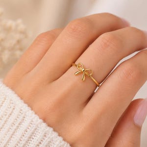 May include: A gold-toned ring featuring a small, centered bow design. The band is thin and polished, with the bow element positioned at the top. The metal has a smooth, reflective finish, creating a simple and clean appearance on the finger.