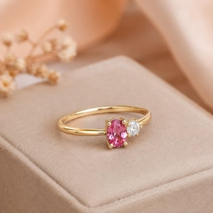 Puede incluir: Un anillo de oro con una piedra preciosa rosa de corte ovalado y una piedra transparente más pequeña de corte redondo, engastadas una al lado de la otra en una banda fina de oro pulido. El anillo se presenta sobre una superficie de tono neutro.