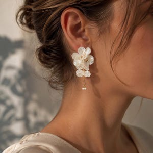 Op de afbeelding: Een witte bloemoorbel met gelaagde, doorschijnende bloemblaadjes in een tros. Een dunne goudkleurige ketting hangt aan de basis van de bloem en eindigt met twee kleine, ronde witte kralen die verticaal aan de hoofdstructuur hangen.