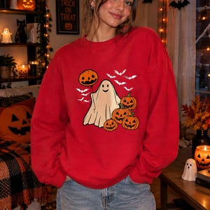 Puede incluir: Una sudadera roja de cuello redondo con un estampado gráfico de un fantasma sonriente que sostiene un globo de calabaza, rodeado de varias calabazas naranjas y pequeños murciélagos blancos. La sudadera tiene mangas largas y cuello acanalado.