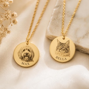 Puede incluir: Dos collares con colgantes circulares dorados en cadenas finas. Un colgante presenta el retrato grabado de un perro con el nombre MAX debajo. El segundo colgante presenta el retrato grabado de un gato con el nombre BELLA debajo.