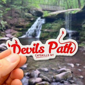 May include: A die-cut vinyl sticker featuring the red text Devils Path in a stylized script font with a small devil tail accent on the letter h and a horn on the letter D, with the red text CATSKILLS NY printed in a simple sans-serif font below.