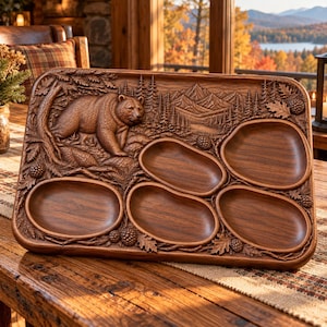 May include: A rectangular wooden serving tray featuring a detailed relief carving of a bear in a forest setting with mountains in the background. The tray includes five smooth, recessed oval compartments for food, surrounded by carved pinecones and foliage.
