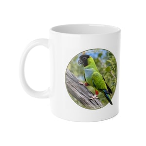 Könnte beinhalten: Ein weißer Keramikbecher mit einer kreisförmigen Grafik eines Nandaysittichs, der auf einem Ast sitzt. Der Vogel hat einen schwarzen Kopf, einen grünen Körper und blau gefärbte Flügelspitzen. Der Text "Nanday Conure" und "Paradise Bird Gifts" ist aufgedruckt.