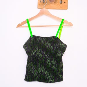 Op de afbeelding: Een zwarte camisole-top met een herhalend patroon van kleine groene digitale tekens en symbolen over de stof. Het kledingstuk heeft een rechte halslijn en twee dunne, fel neongroene spaghettibandjes, hangend aan een houten kledinghanger.