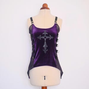 May include: A sleeveless purple velvet top featuring a black cross graphic on the front. The garment has black side panels with silver-toned metal O-rings and straps, a scoop neckline, and a high-low hemline displayed on a dress form.