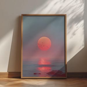 May include: A framed vertical wall art print depicting a large, vibrant red sun setting over a calm ocean horizon. A small flock of birds is silhouetted against the sun, with its reflection shimmering on the water surface and rocks in the foreground.