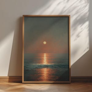 May include: A rectangular wall art print in a light wood frame featuring a seascape at sunset. The sun is a small, pale yellow circle centred in a dark, hazy sky above a calm ocean. A bright, vertical reflection of the sun glows on the water.